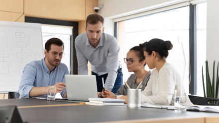 Four colleagues gather around a laptop in a meeting room, one man standing and pointing at the screen while others look on