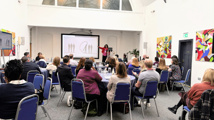Audience seated at round tables listening to a speaker presenting beside a screen at a professional forum in a bright conference room.