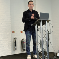Male presenter standing behind a lectern with a laptop, speaking to an audience during a conference session.