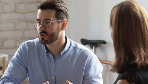 Man wearing glasses speaks and gestures during a discussion with a colleague in a bright office setting.