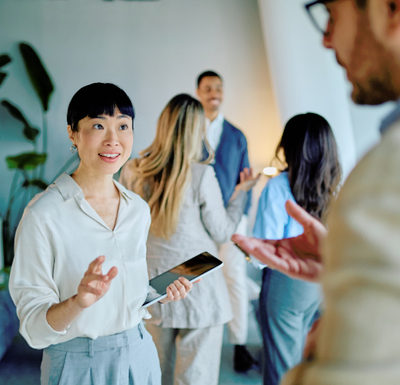 Woman holding a smartphone and gesturing while speaking to a colleague in a bright office, with a small group talking in the background.
