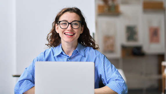 Smiling woman wearing glasses sits at a desk working on a laptop in a modern workspace.