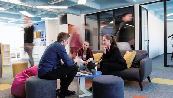 Three colleagues seated in a modern office lounge discuss work around a laptop, while other people move quickly in the background.