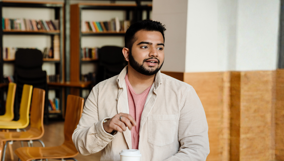 Man in a casual shirt holds a coffee cup while speaking in a relaxed office or breakout area with chairs and bookshelves behind him.