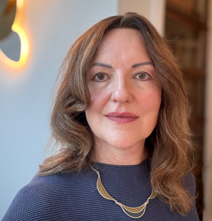 Portrait photo of Liz Reynolds. Liz has wavy auburn shoulder length hair and is wearing a blue top and a gold necklace.