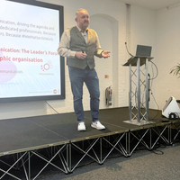Male speaker presenting on stage beside a screen at a conference, gesturing while speaking with a laptop on a lectern.