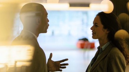 Two colleagues stand facing each other in conversation in a softly lit office setting, seen through glass with warm reflections and blurred lights 